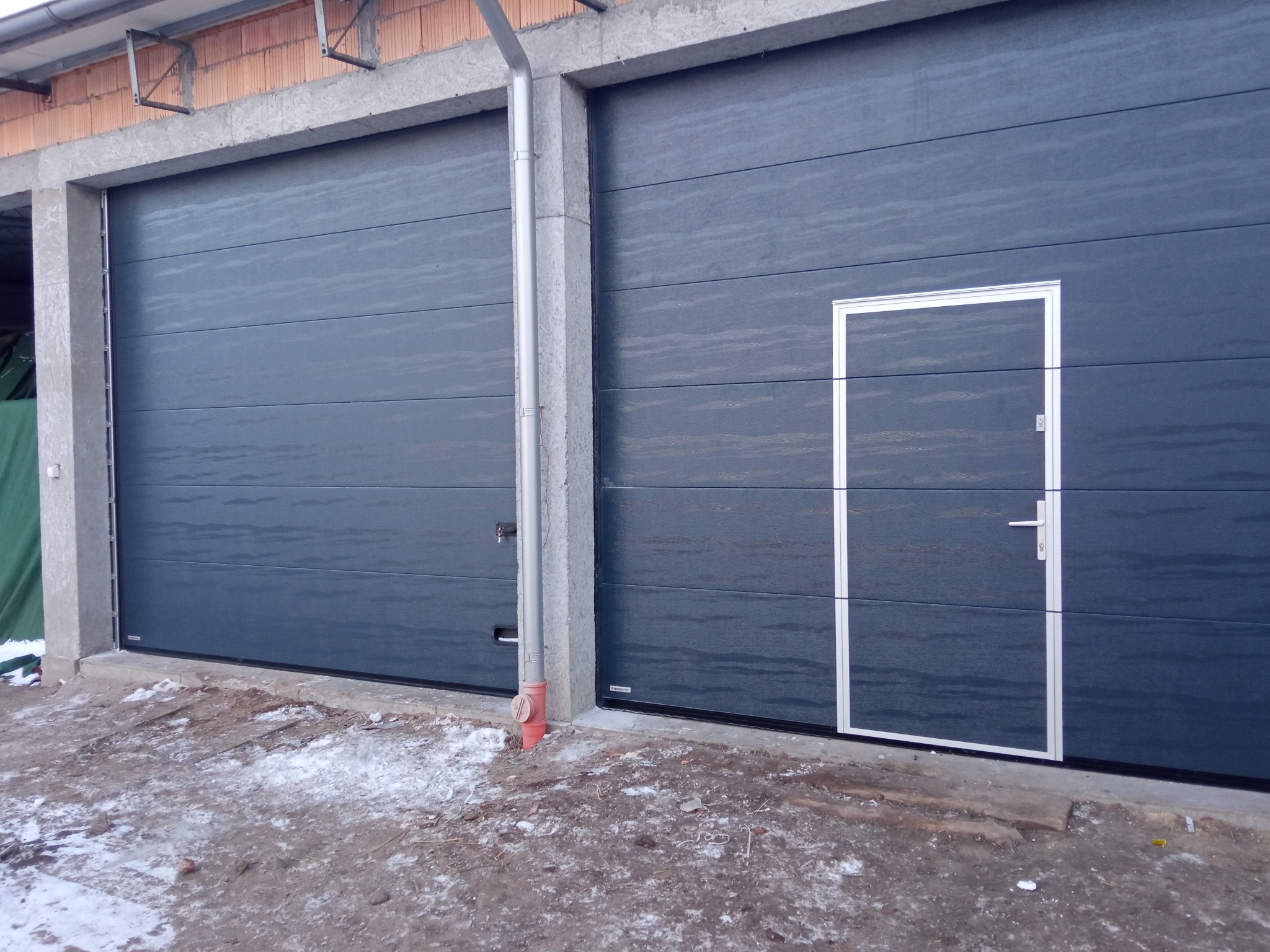Dwie szare segmentowe bramy garażowe, jedna z wbudowanymi drzwiami z białym obramowaniem i klamką, na zewnątrz resztki śniegu.
