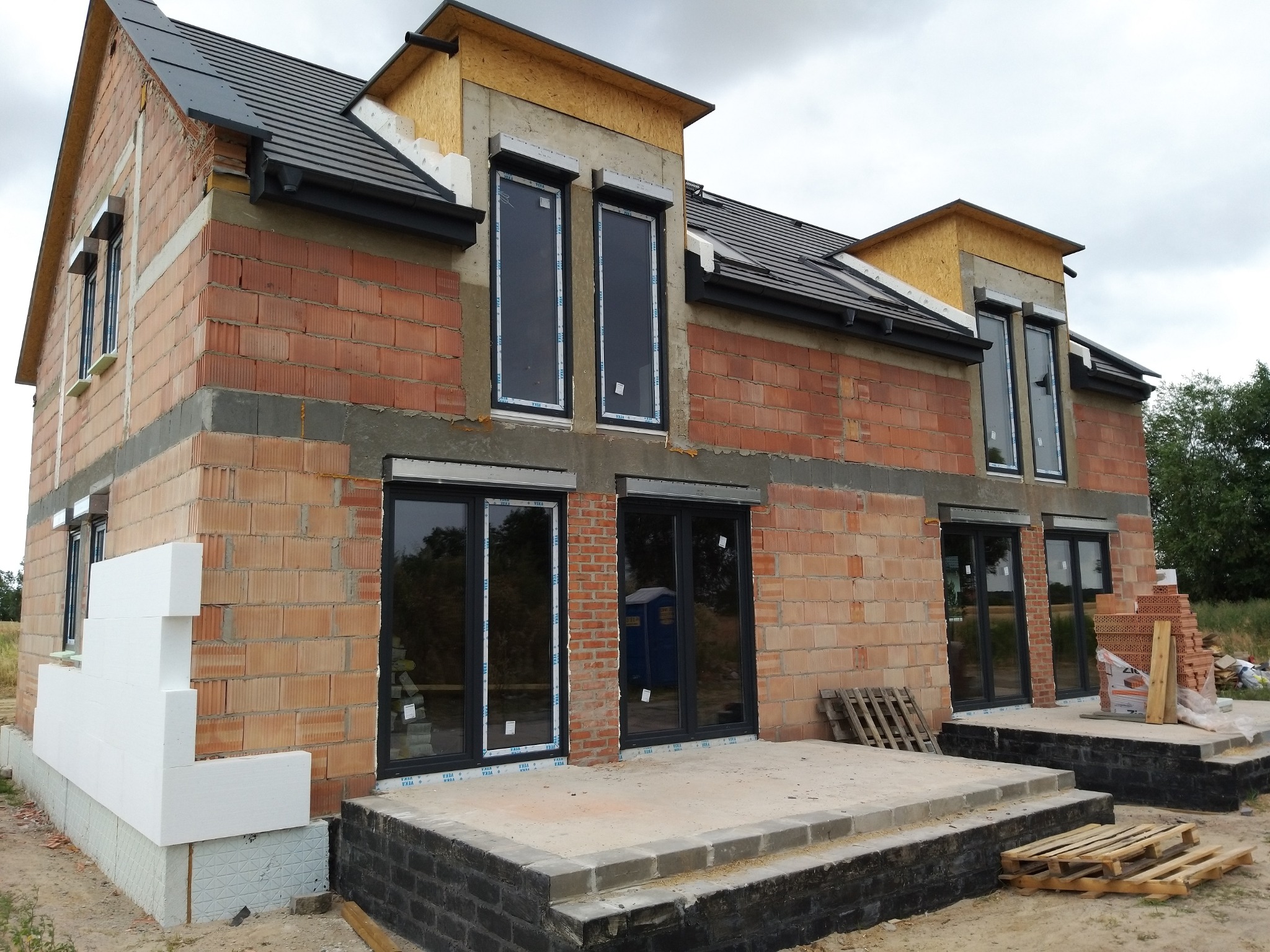 Surowy stan budynku jednorodzinnego z zamontowanymi oknami w czarnych ramach, widoczne ocieplenie styropianem na fragmentach elewacji oraz niezabezpieczone rolety zewnętrzne w górnej części okien.