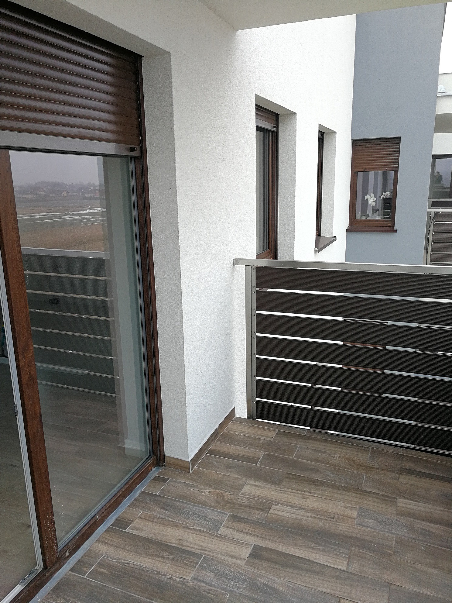 Zewnętrzny widok balkonu z brązowymi roletami opuszczonymi na oknach, drewnopodobna podłoga z płytek oraz nowoczesne balustrady balkonowe z poziomych listew.