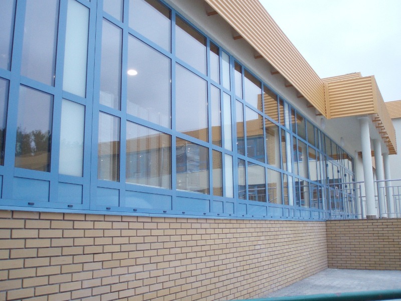 Długi rząd okien PCV z niebieskimi ramami w budynku w Poznaniu, widoczny z zewnątrz, z ceglaną ścianą w dolnej części.