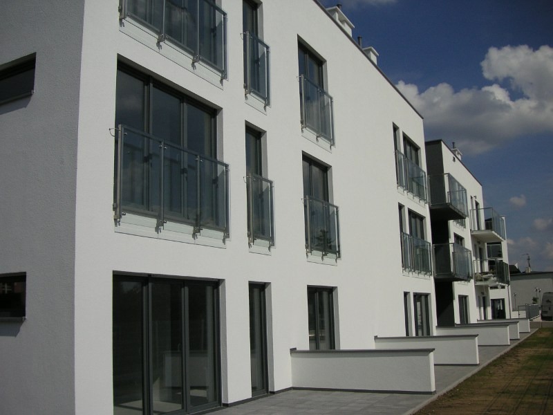 Nowoczesny budynek mieszkalny z białymi ścianami i balkonami z szklanymi balustradami, widok z zewnątrz w słoneczny dzień.