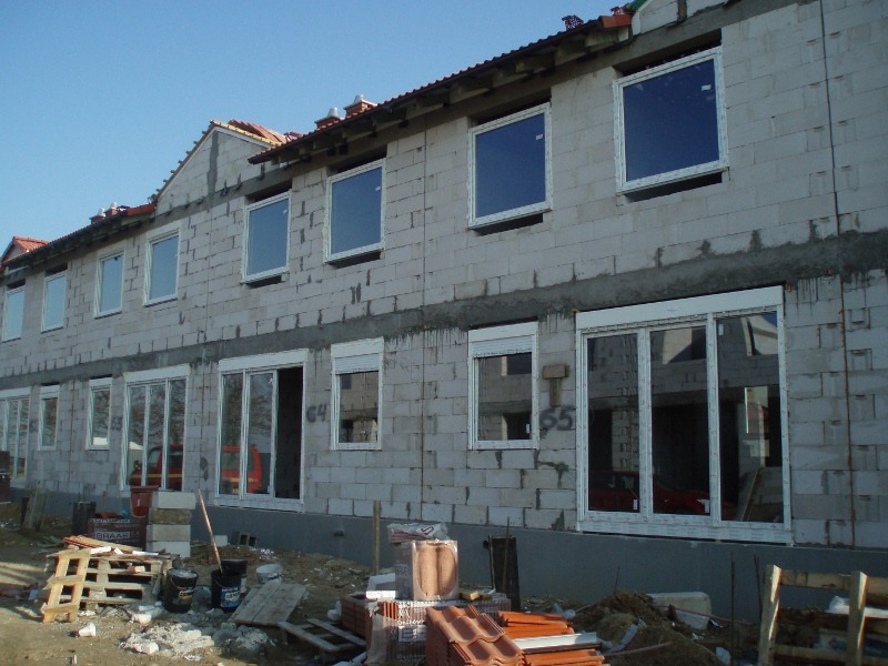 Budynek w trakcie budowy z zamontowanymi białymi oknami i roletami zewnętrznymi, widoczne materiały budowlane i rusztowania.