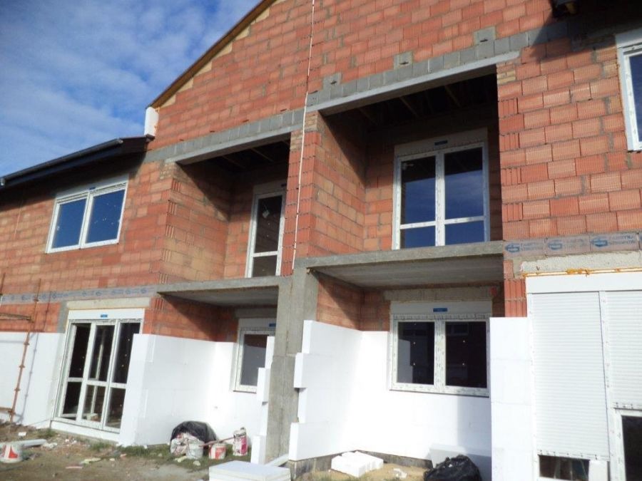 Budynek w trakcie budowy z zamontowanymi oknami z białymi ramami i częściowo zamontowanymi roletami zewnętrznymi, widoczne ocieplenie styropianem.