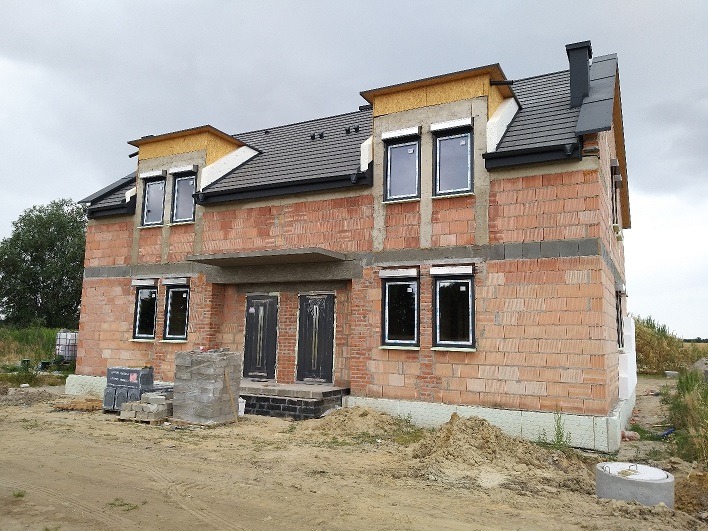 Surowy stan budynku z zamontowanymi oknami z czarnymi ramami i przygotowanymi otworami na rolety zewnętrzne, widoczne materiały budowlane na placu budowy.