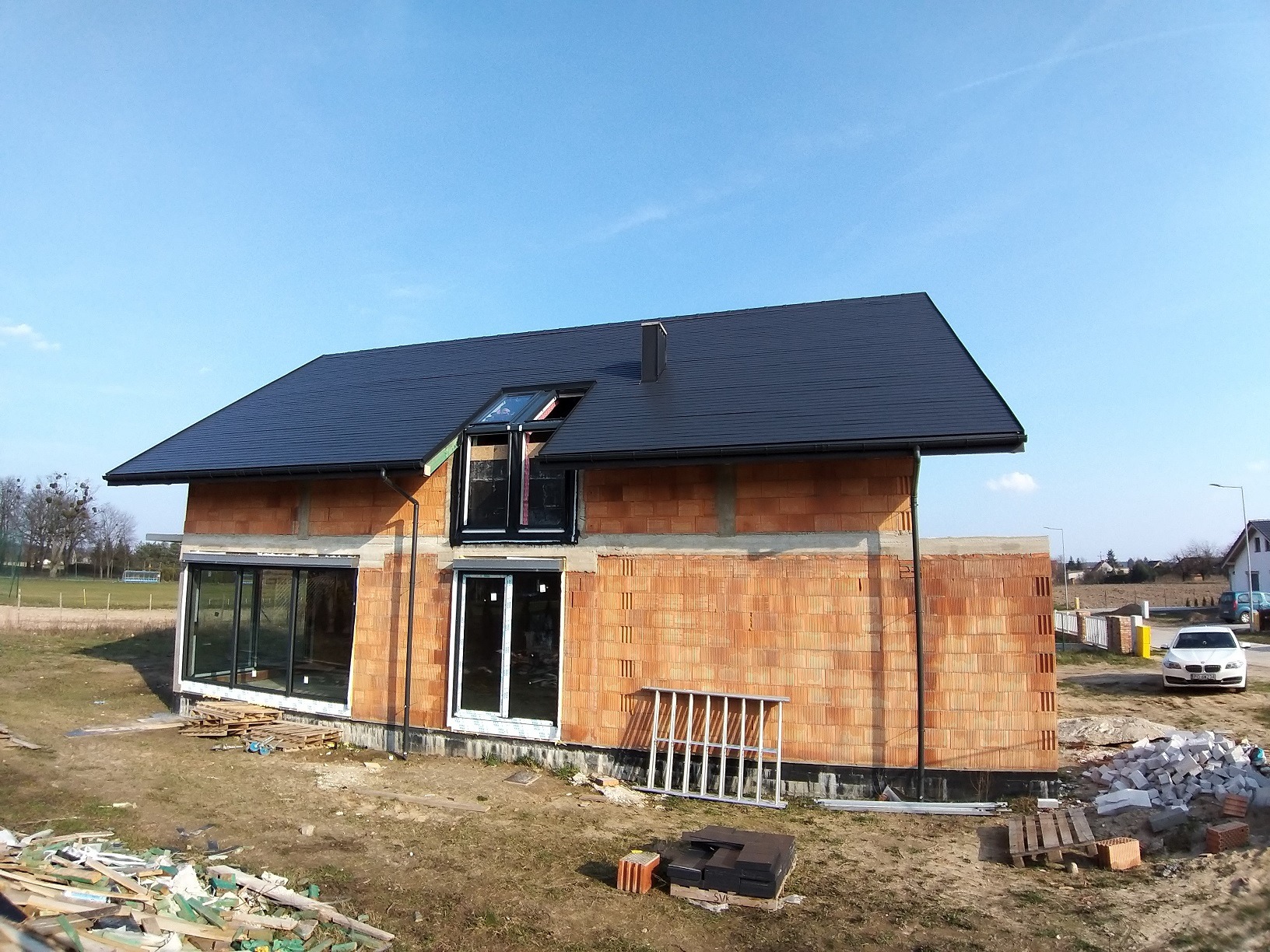 Dom jednorodzinny w trakcie budowy, widoczne wstawione okna z czarnymi ramami i częściowo zamontowane rolety, ceglany mur i czarny dach, na ziemi leżą materiały budowlane.