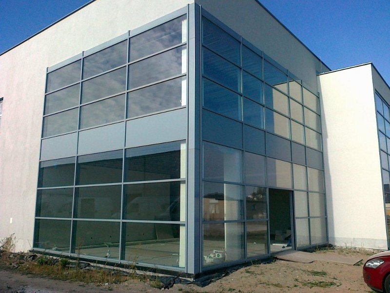 Duża fasada budynku z wieloma szklanymi panelami w szarych ramach, widoczne wnętrze w stanie surowym, ziemia wokół budynku niezagospodarowana, fragment czerwonego samochodu w prawym dolnym rogu.