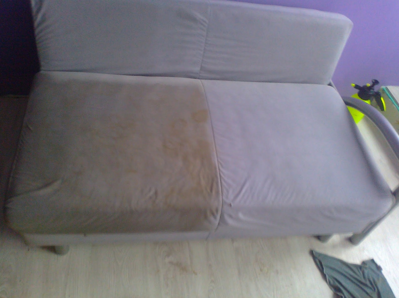 Szara rozkładana sofa z widoczną różnicą w czystości tapicerki: lewa strona brudna z plamami, prawa strona świeżo wyczyszczona, obok leży szara szmatka.