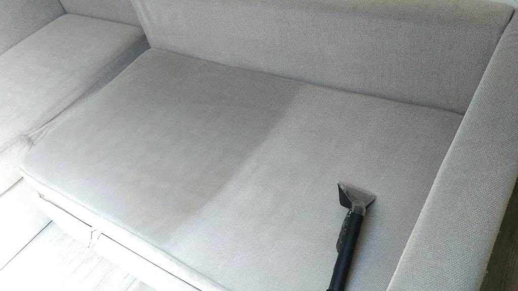 Szara sofa z widoczną różnicą w czystości tapicerki po częściowym czyszczeniu, z dyszą odkurzacza piorącego na powierzchni.