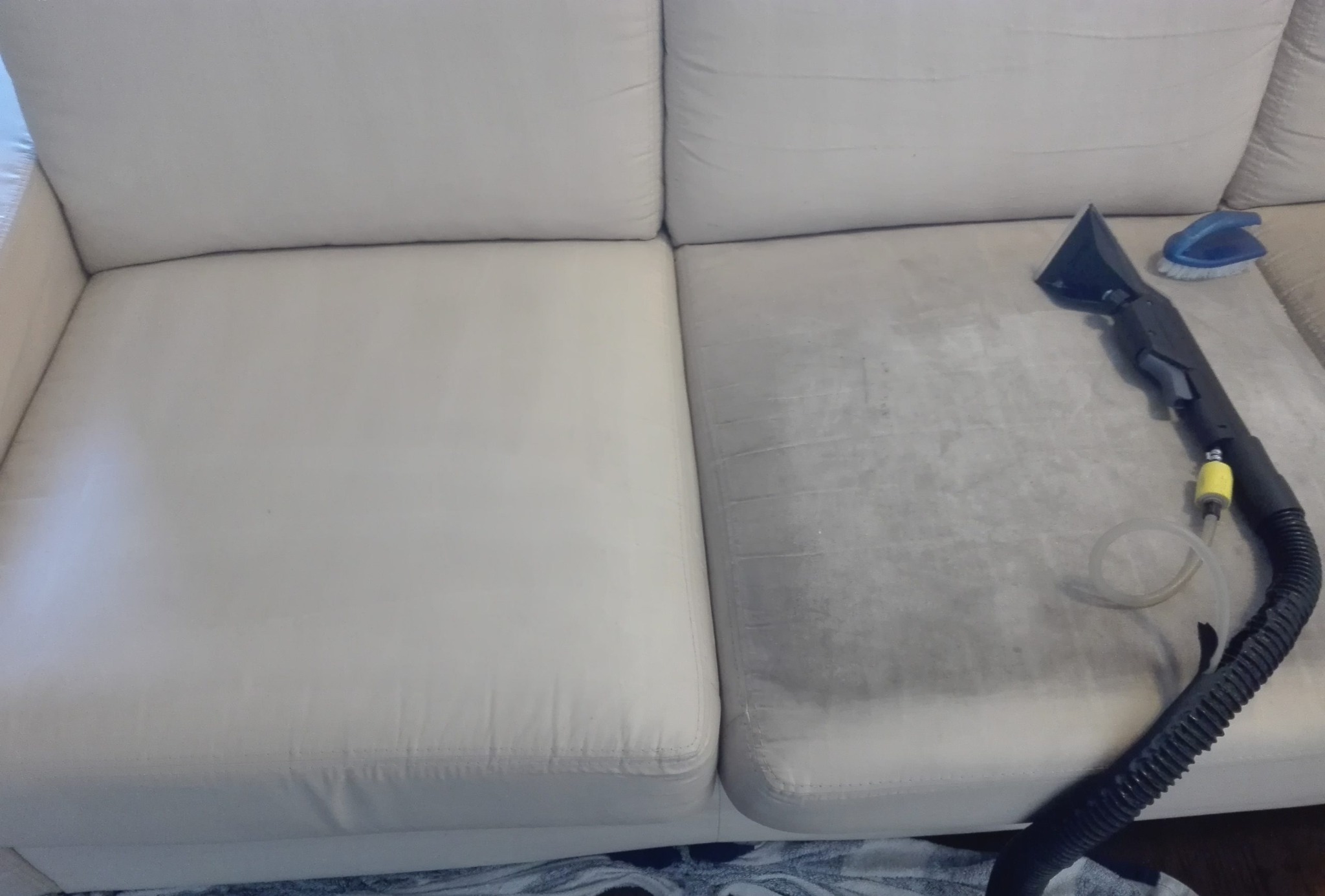 Jasna kanapa z widoczną różnicą czystości pomiędzy wyczyszczoną a nie wyczyszczoną częścią siedziska, obok leży końcówka odkurzacza piorącego i szczotka.