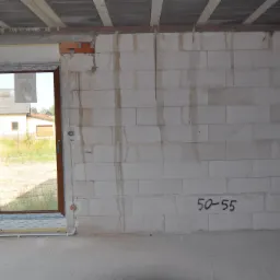 Surowy stan wnętrza z widocznymi przewodami elektrycznymi w ścianach z betonu komórkowego, okna z brązowymi ramami, widok na zewnątrz z zielenią.