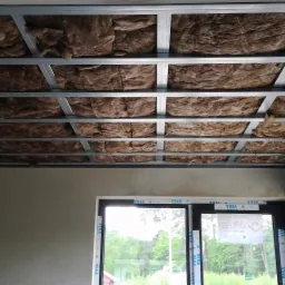 Sufit podwieszany z metalową konstrukcją wypełnioną brązową wełną mineralną, widok z dołu, okno z widokiem na zieleń w tle.