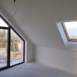 Poddasze po gładziowaniu i malowaniu na biało, widoczne okno dachowe, duże okno z widokiem na ogród i przygotowane punkty elektryczne na ścianie.