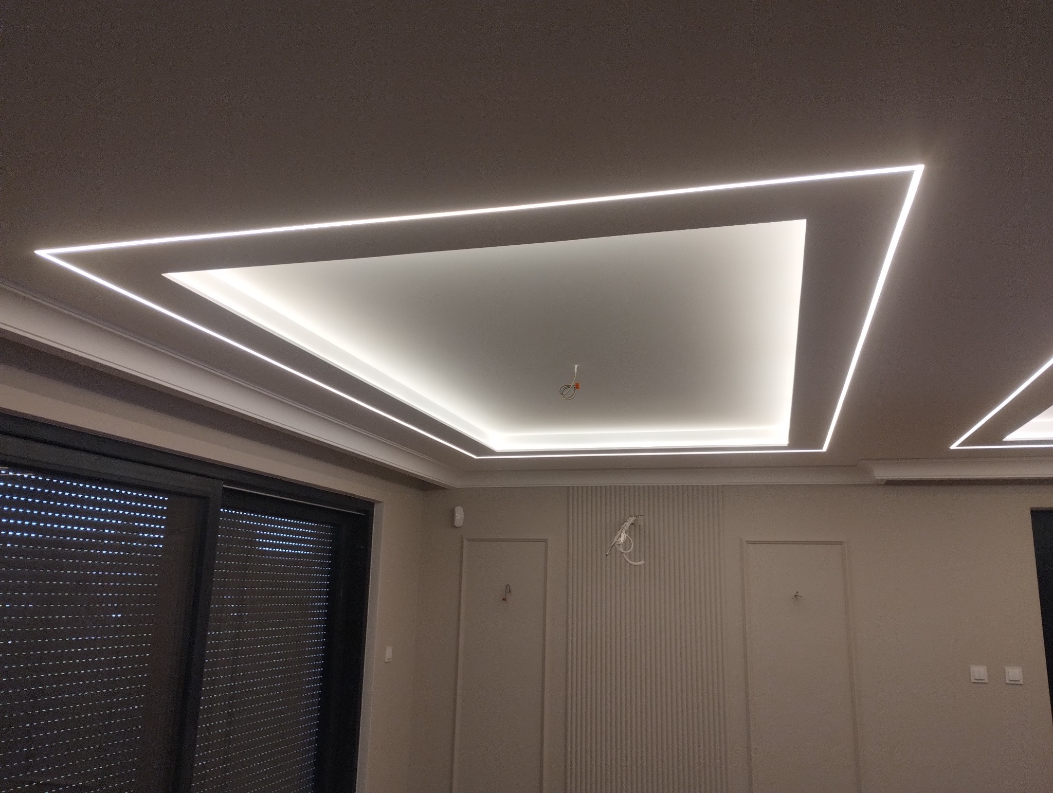 Nowoczesny sufit podwieszany z oświetleniem LED w kształcie kwadratu, wykończony listwami dekoracyjnymi. Widoczne okno z żaluzjami i minimalistyczne drzwi.