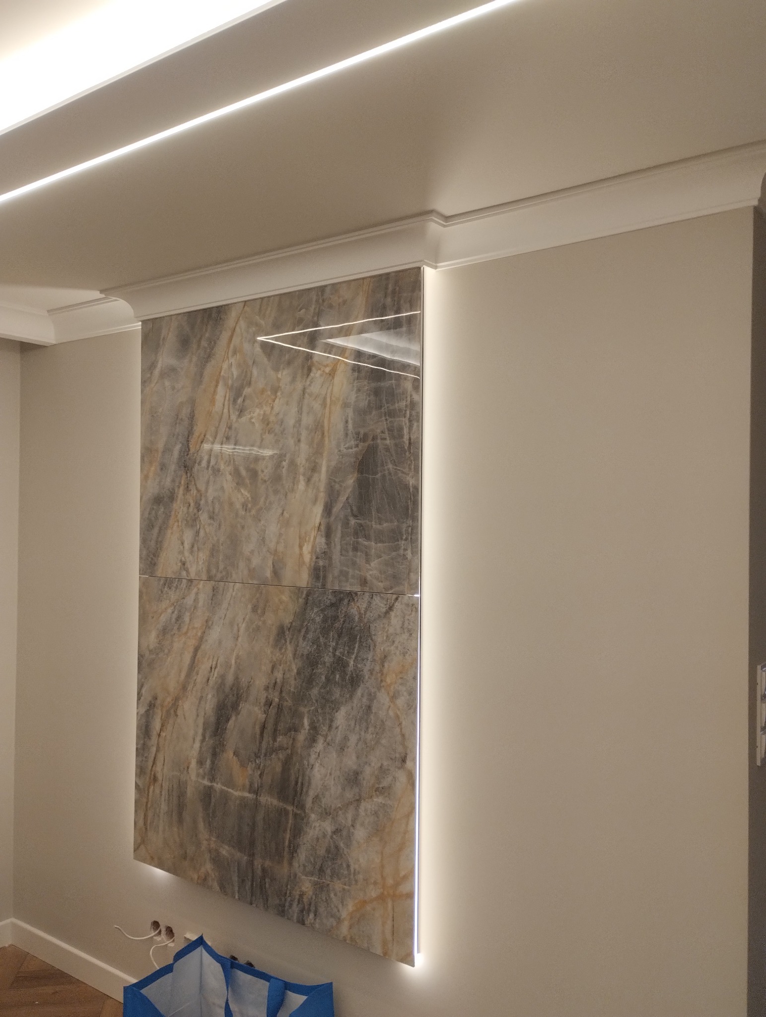 Ściana z płyt imitujących marmur, podświetlona ledami. Dekoracyjne listwy sufitowe. Estetyczne wykończenie wnętrza z elementami oświetlenia.