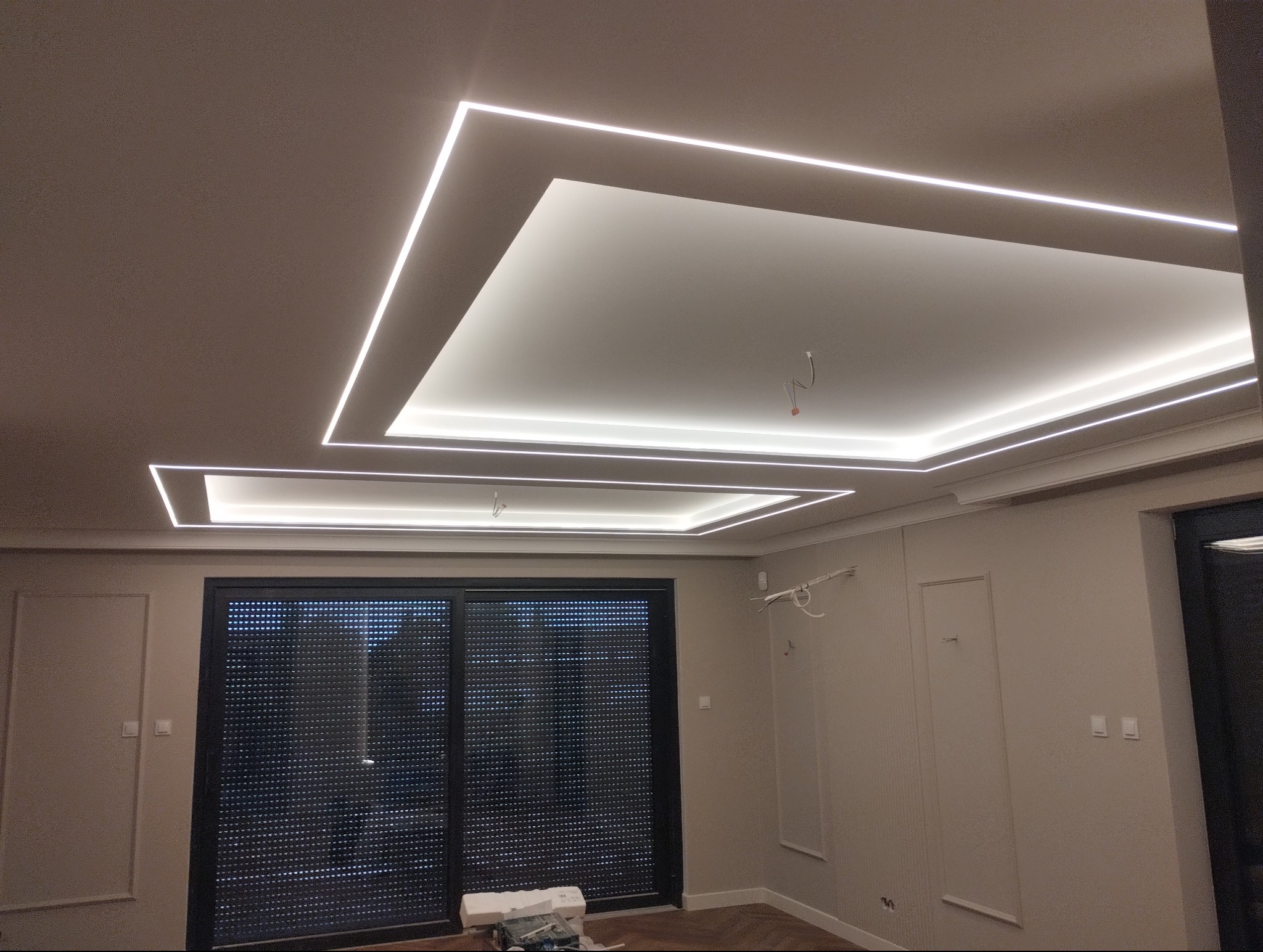 Nowoczesne oświetlenie sufitowe LED w kształcie prostokątów, podwieszany sufit,  w wykończonym wnętrzu. Widoczne okno z roletami zewnętrznymi i fragment ściany.