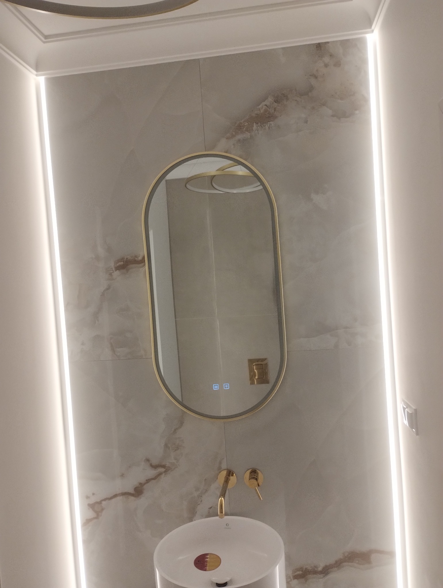 Elegancka łazienka z owalnym lustrem z podświetleniem LED, armaturą w kolorze złota i marmurową okładziną ściany. Nowoczesny design i dbałość o detale.