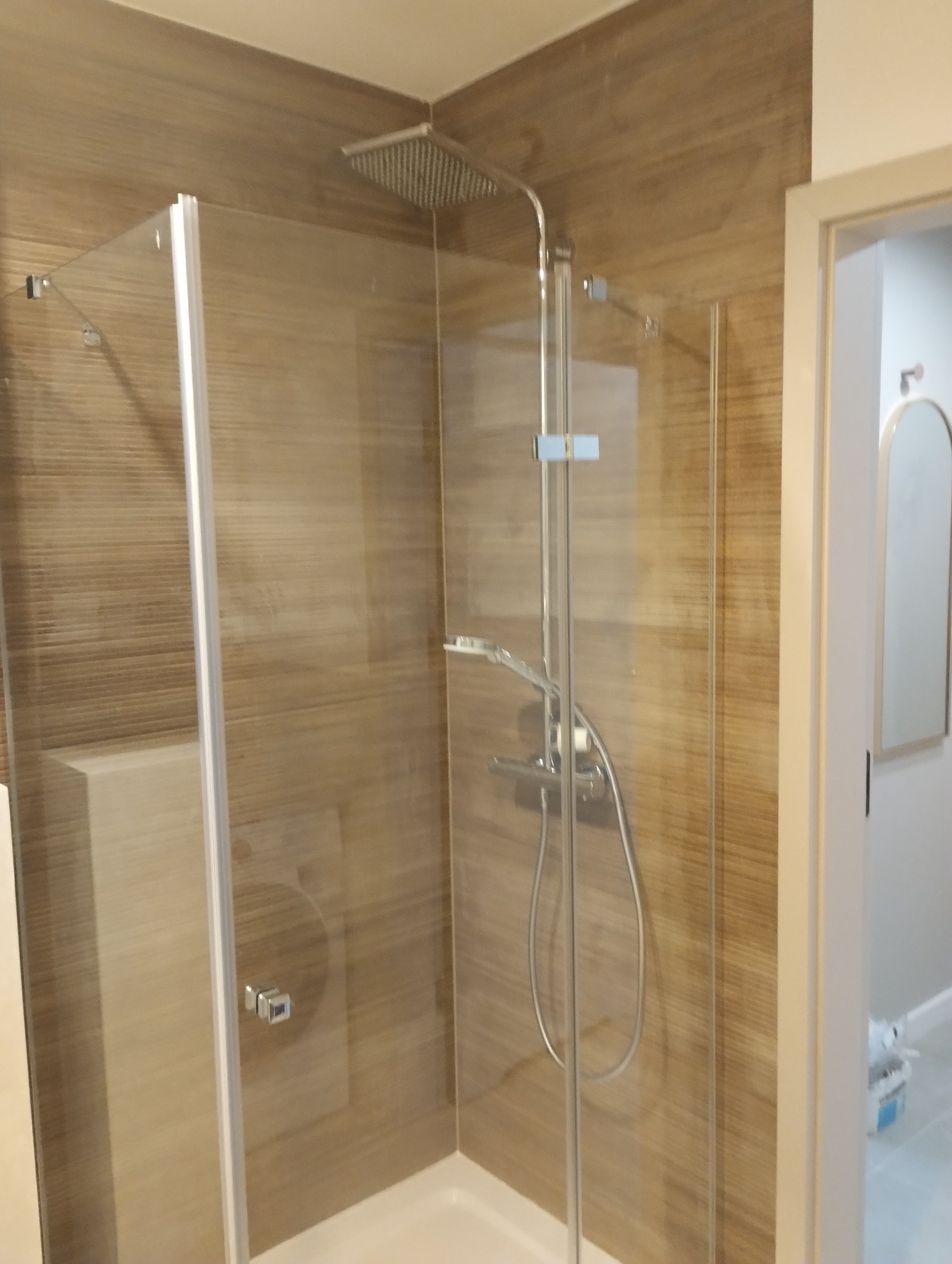 Kabina prysznicowa z deszczownicą i brązowymi płytkami imitującymi drewno. Widoczny fragment lustra i drzwi do łazienki.