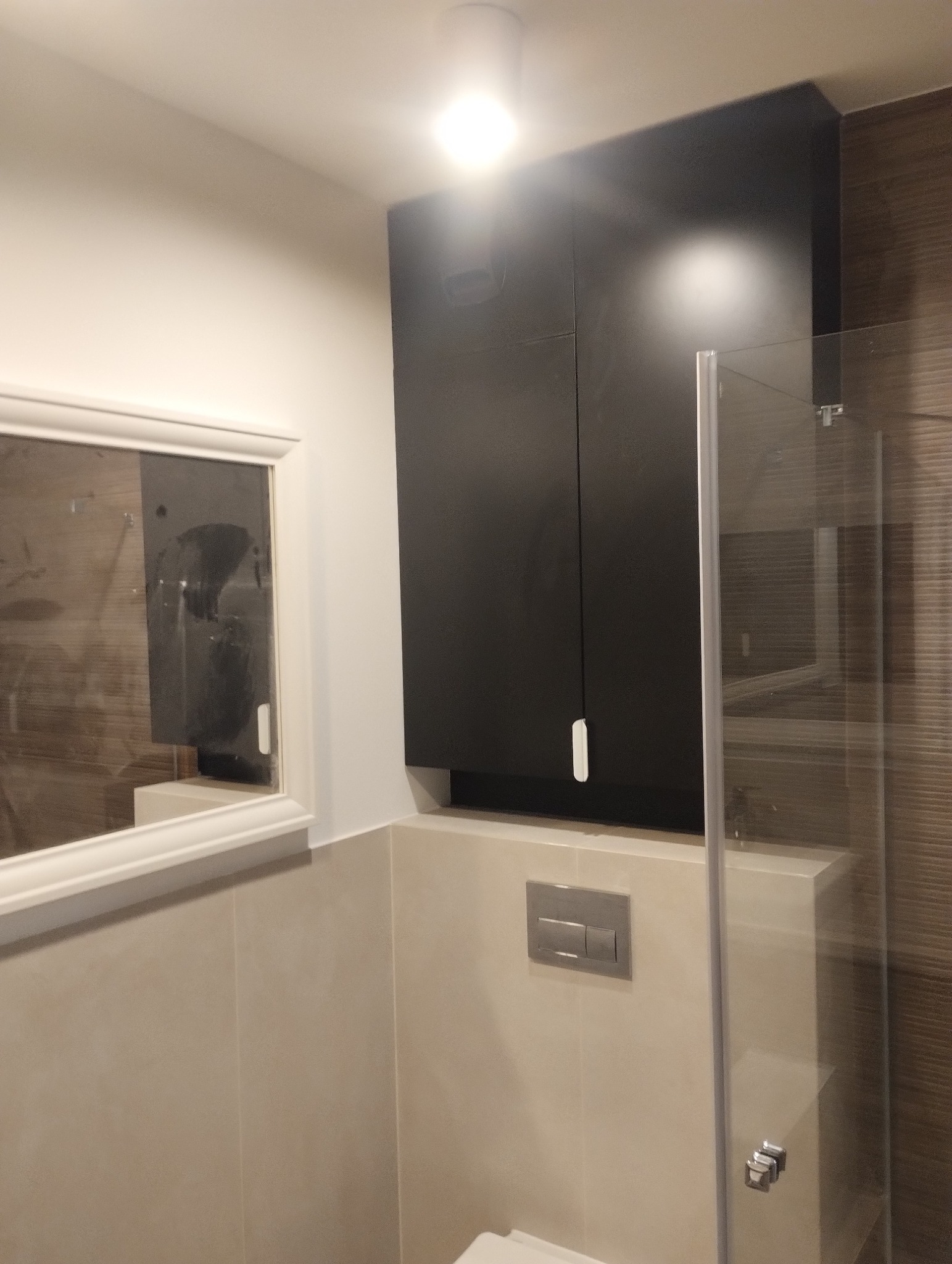 Nowoczesna łazienka z ciemną szafką nad zabudowaną spłuczką WC, lustrem w białej ramie i szklaną kabiną prysznicową. Jasne płytki na ścianach.