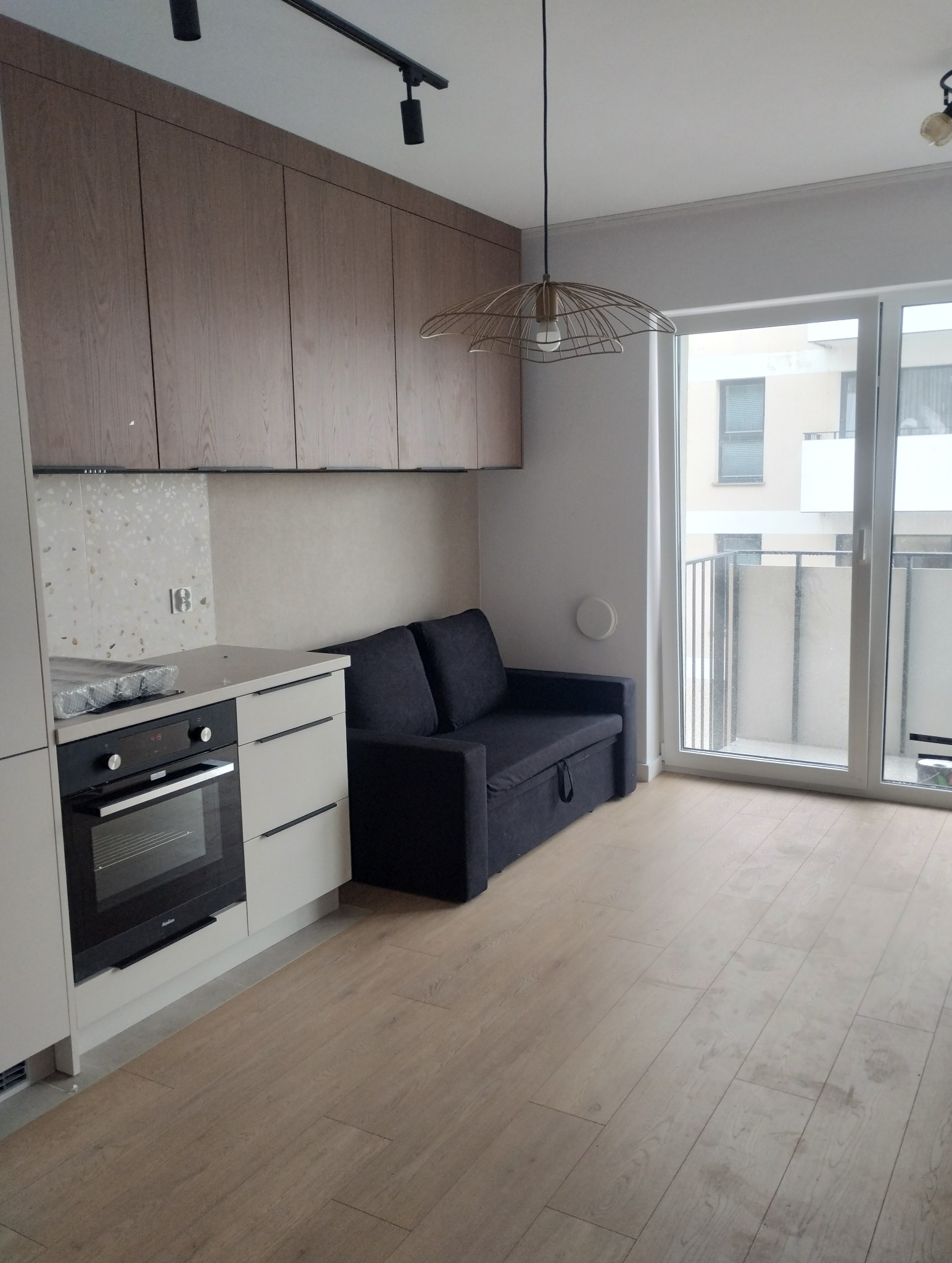 Jasne wnętrze: zabudowa kuchenna z piekarnikiem, szara sofa pod szafkami, drewniana podłoga i lampa w stylu industrialnym. Balkon z widokiem na budynek.