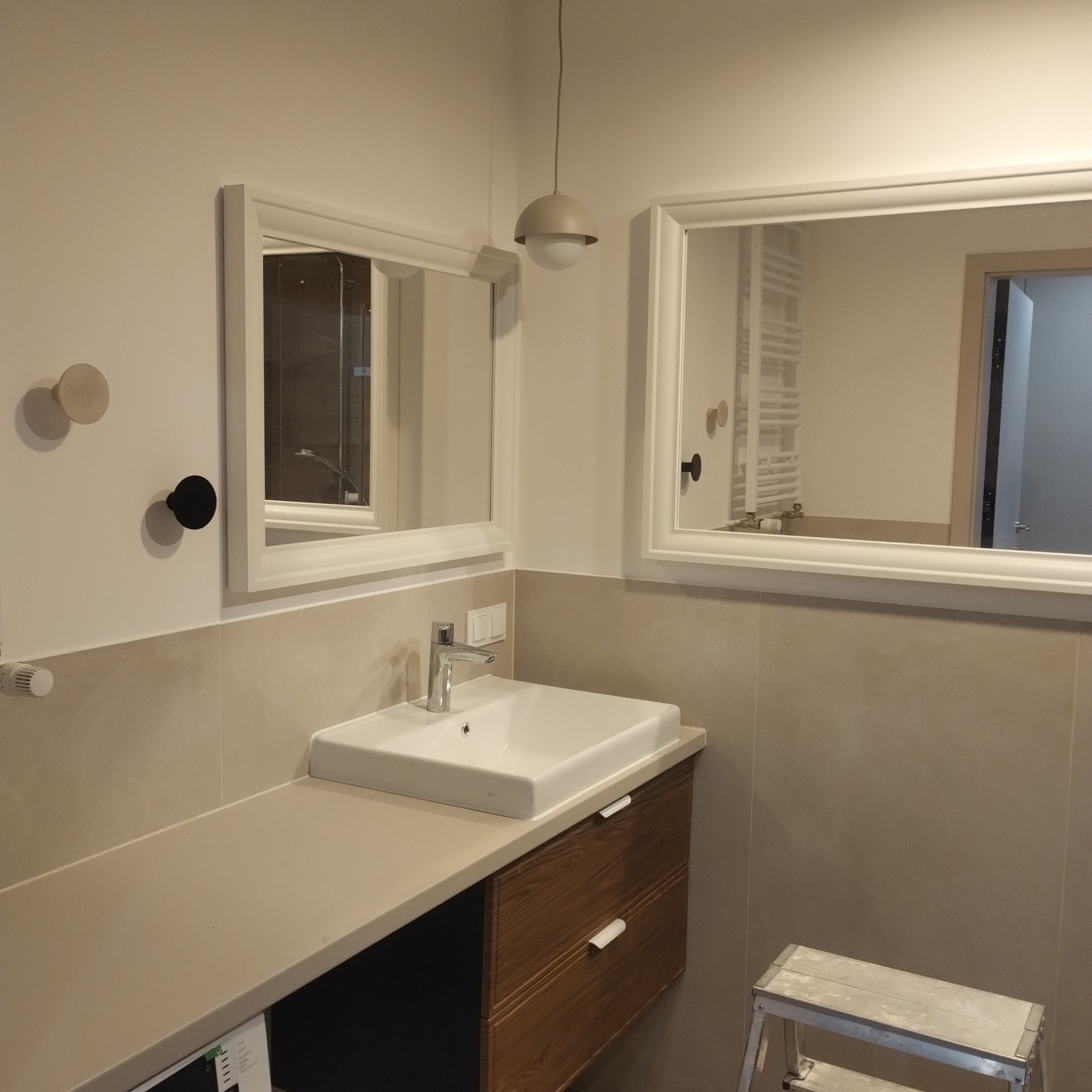 Nowoczesna łazienka po remoncie: biała umywalka nablatowa, drewniana szafka, dwa lustra w białych ramach i minimalistyczna lampa wisząca.