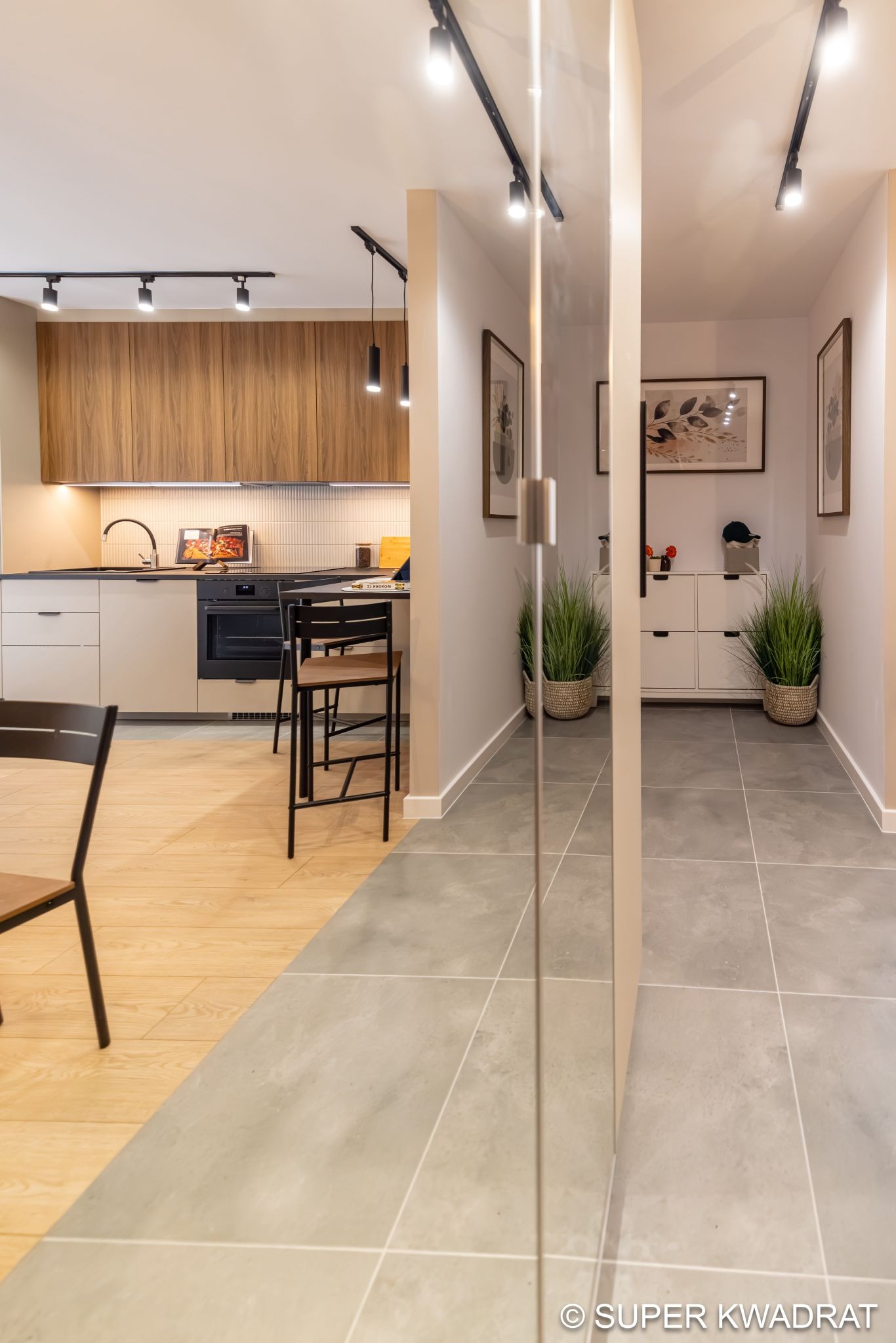 Nowoczesne wnętrze: kuchnia z drewnianymi szafkami i jasną podłogą przechodzi w przedpokój z szarą podłogą z płytek, oddzielone szklanymi drzwiami.