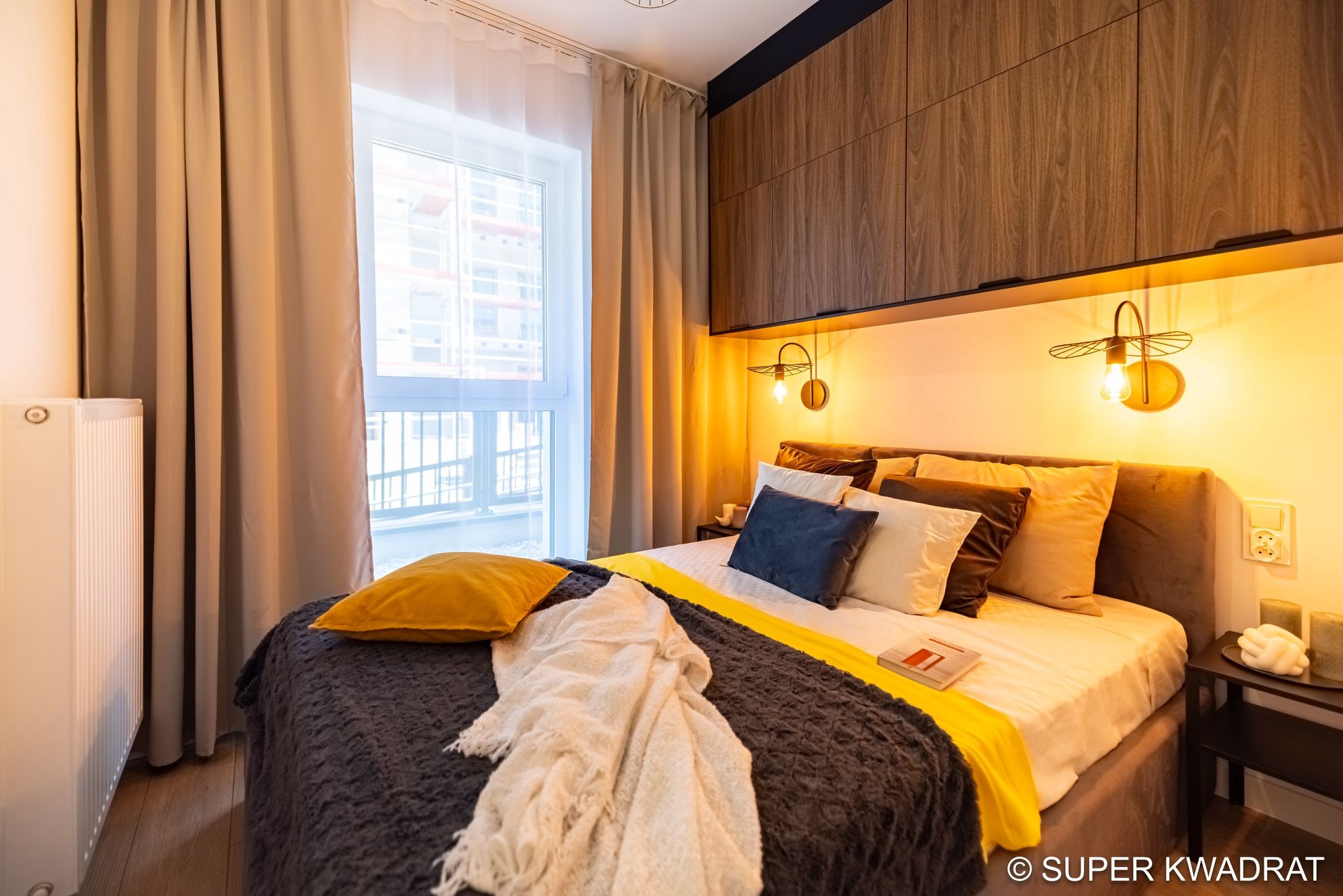 Przytulna sypialnia z drewnianymi szafkami nad łóżkiem, oświetlona ciepłym światłem kinkietów. Okno z jasnymi zasłonami w tle. Dekoracyjne poduszki i pled na łóżku.