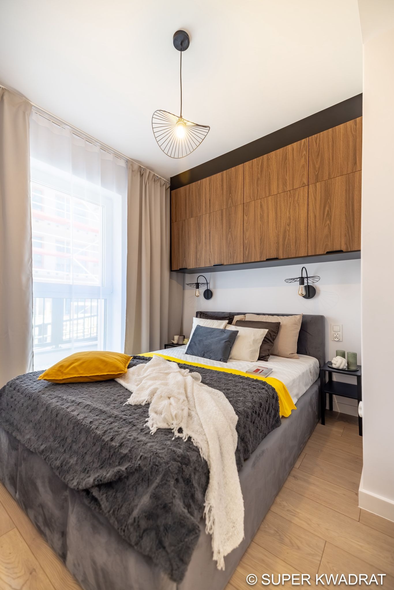 Nowoczesna sypialnia z drewnianą zabudową nad łóżkiem, lampkami ściennymi i żółtymi akcentami kolorystycznymi. Widoczne okno z zasłonami i stolik nocny.