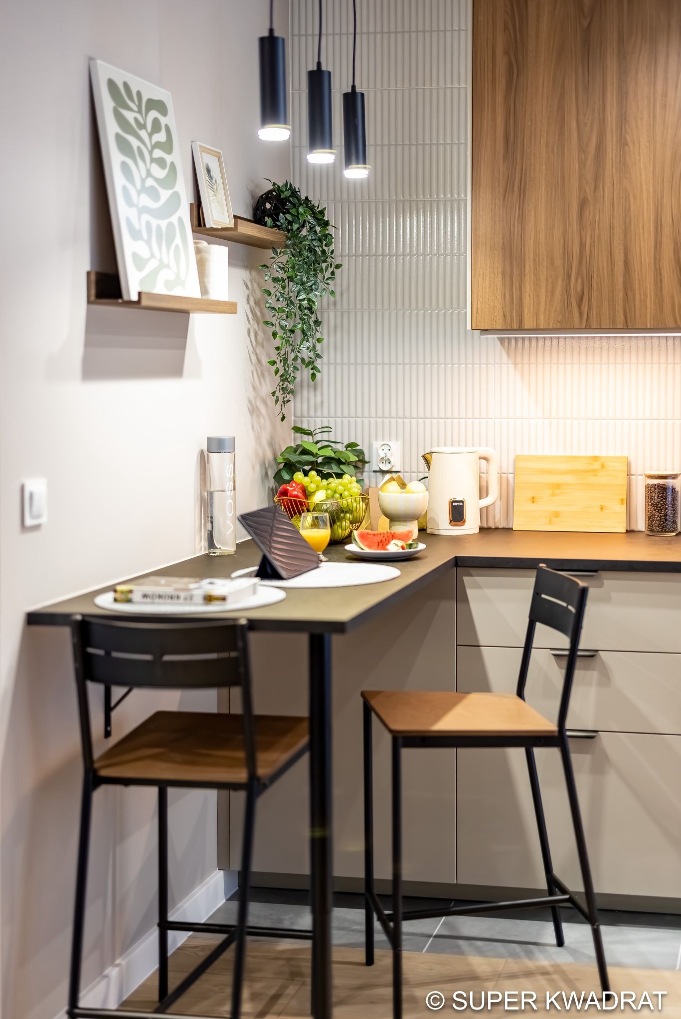 Nowoczesna kuchnia z barkiem: płytki imitujące fale, drewniane szafki, czarne lampy wiszące. Na blacie owoce, napoje i dekoracje. Dwa krzesła barowe.