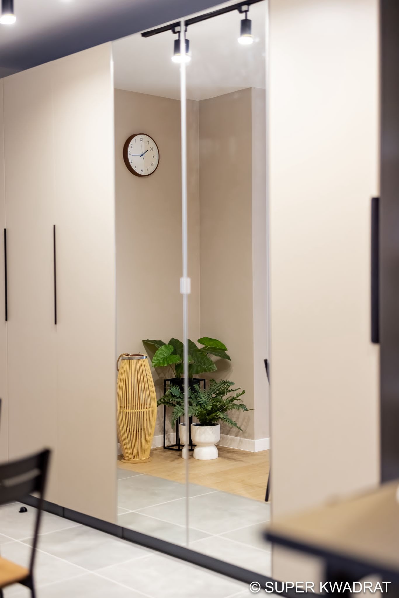 Beżowa szafa wnękowa z lustrzanymi drzwiami przesuwnymi, odbijającymi wnętrze z zegarem, roślinami i drewnianą podłogą. Minimalistyczny design, neutralna kolorystyka.