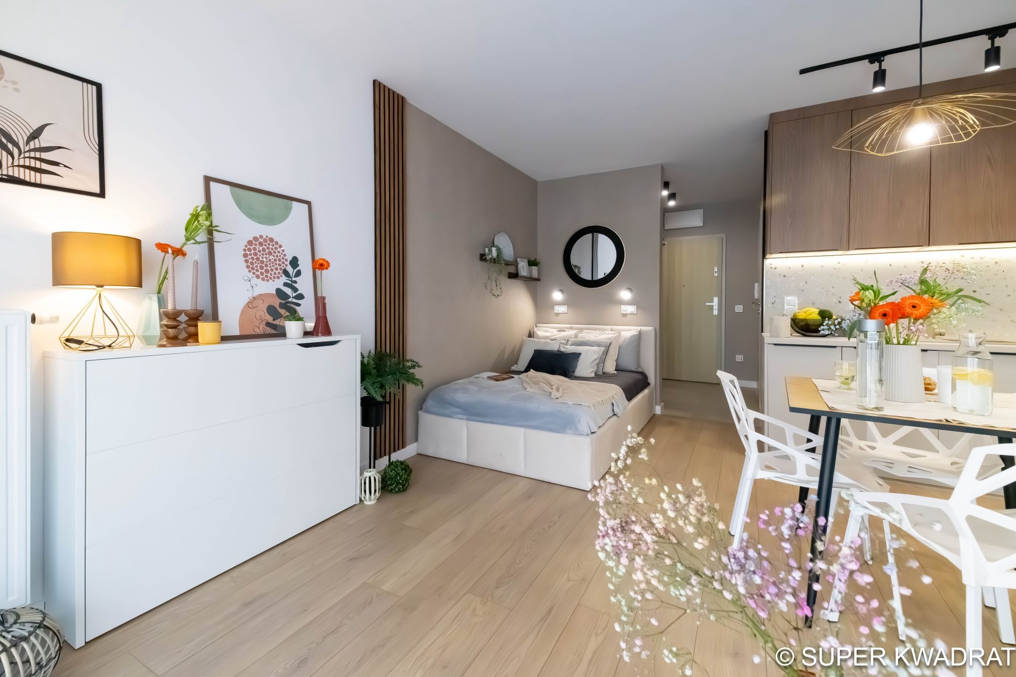 Jasne, nowocześnie urządzone studio: drewniana podłoga, białe meble, łóżko, stół z krzesłami i dekoracje roślinne. Minimalistyczny design z akcentami kolorystycznymi.