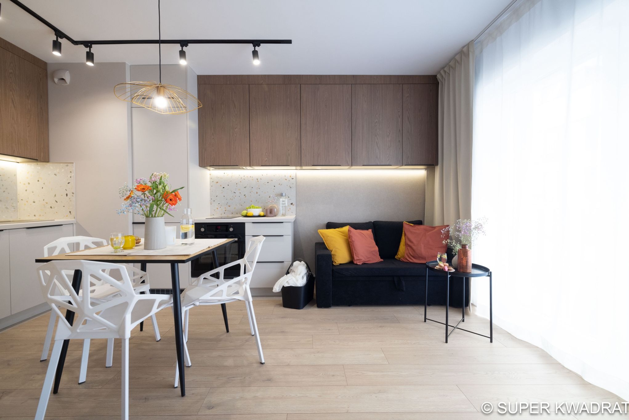 Jasne wnętrze mieszkania z drewnianą podłogą, stołem z kwiatami i sofą z poduszkami. Widoczne szafki kuchenne i oświetlenie punktowe.