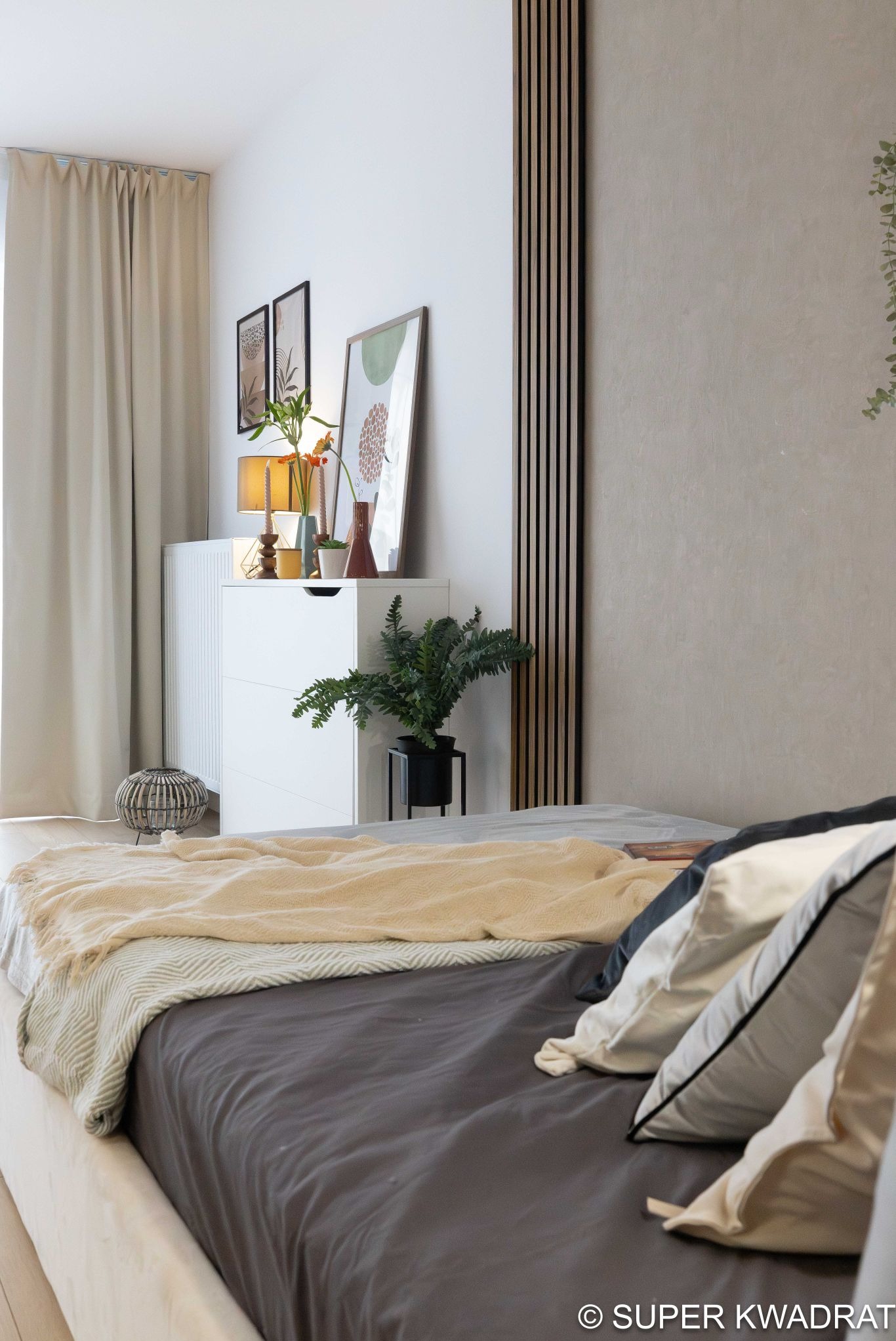 Jasna sypialnia z łóżkiem, komodą i drewnianymi panelami ściennymi. Dekoracje roślinne i minimalistyczne obrazy dodają wnętrzu przytulności.