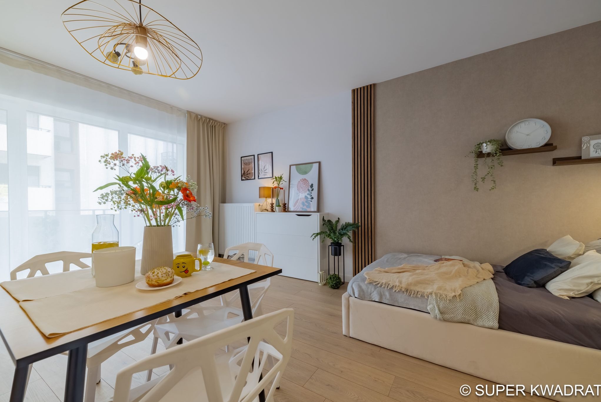 Jasne wnętrze pokoju z drewnianą podłogą, stołem jadalnym z kwiatami i łóżkiem. Dekoracyjne panele ścienne i minimalistyczne dodatki.