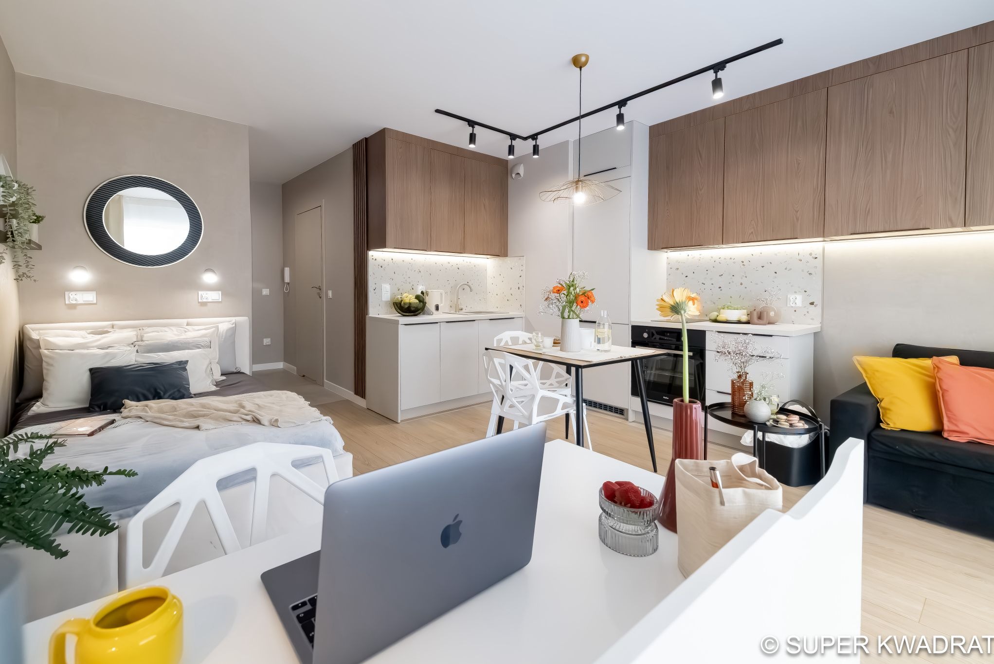 Nowoczesne studio: jasne ściany, drewniane szafki kuchenne, okrągłe lustro nad łóżkiem, designerskie oświetlenie i laptop na biurku. Minimalistyczny, funkcjonalny design.