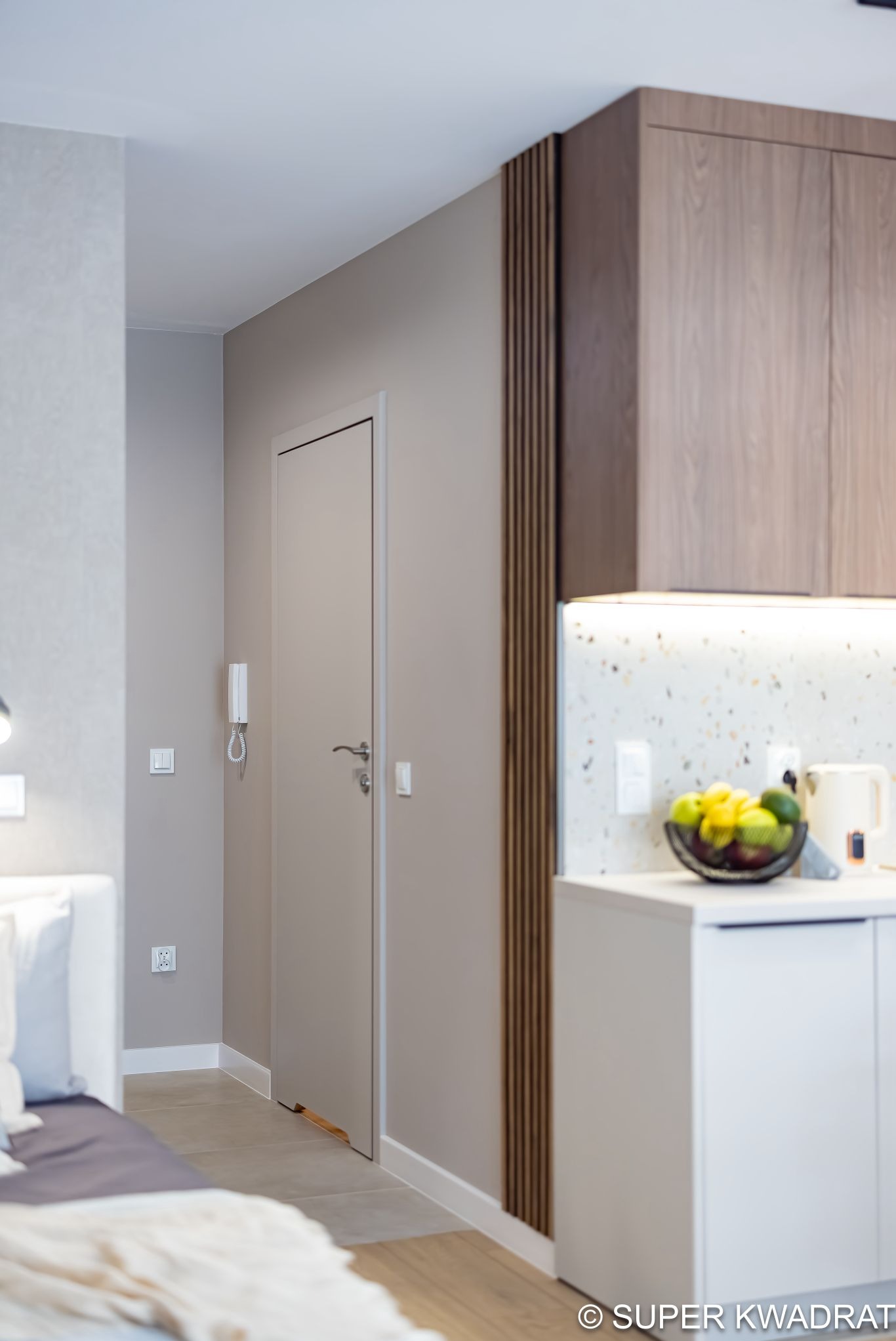 Nowoczesne wykończenie wnętrza: szara ściana, białe listwy przypodłogowe, drewniane lamele dekoracyjne, zabudowa kuchenna z owocami i czajnikiem, widok z perspektywy łóżka.