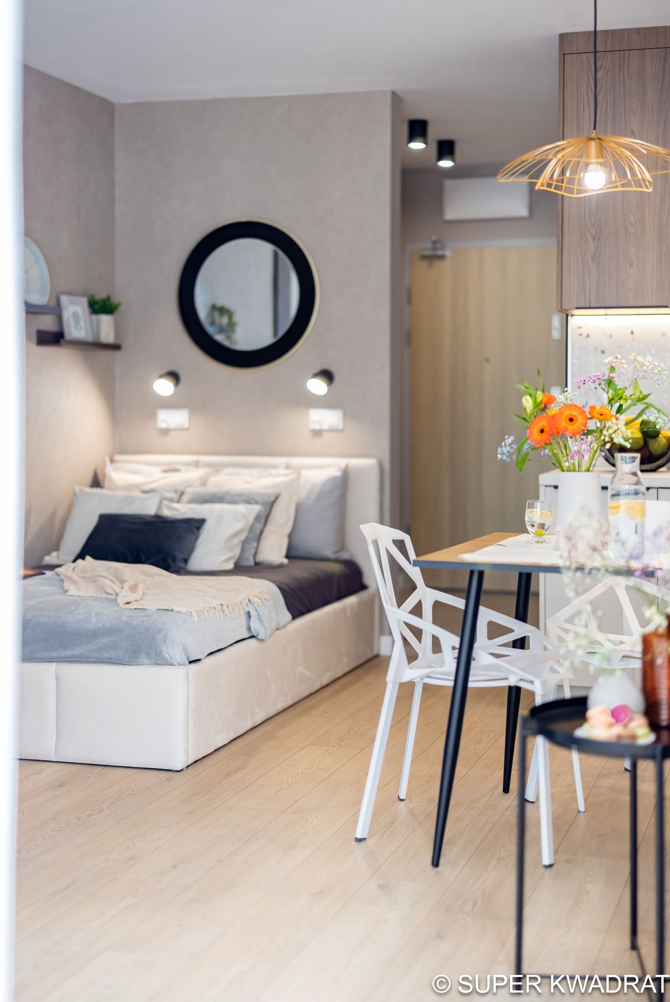 Jasne wnętrze mieszkania z łóżkiem, okrągłym lustrem i stołem z kwiatami. Nowoczesny design, neutralna kolorystyka, drewniana podłoga. Oświetlenie punktowe.
