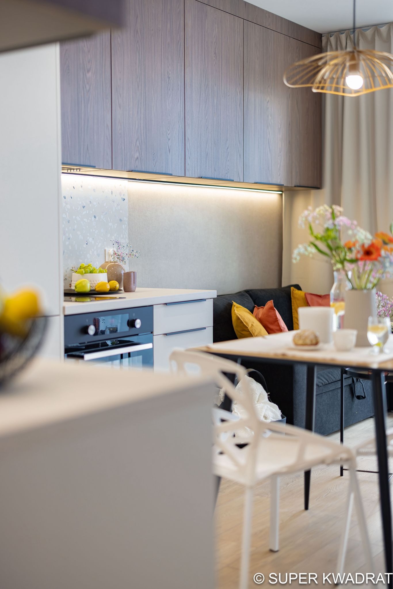 Nowoczesna kuchnia z drewnianymi szafkami, oświetleniem LED pod szafkami i blatem z owocami. W tle salon z sofą i kwiatami na stole. Całość w jasnych, stonowanych barwach.