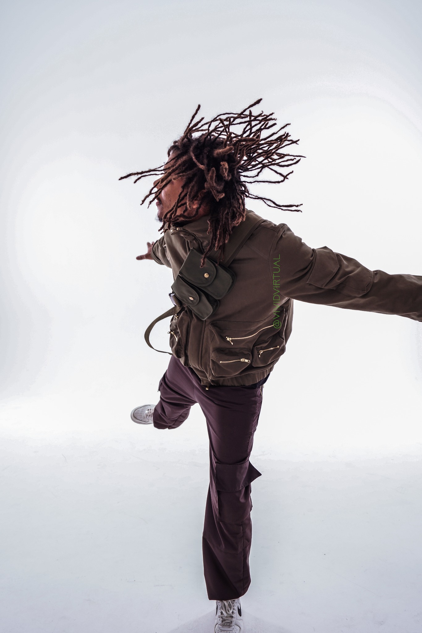 Mężczyzna z dredami w brązowej kurtce i spodniach cargo, w ruchu, na białym tle. Widoczny fragment nazwy firmy @VIVIDVIRTUAL na kurtce.