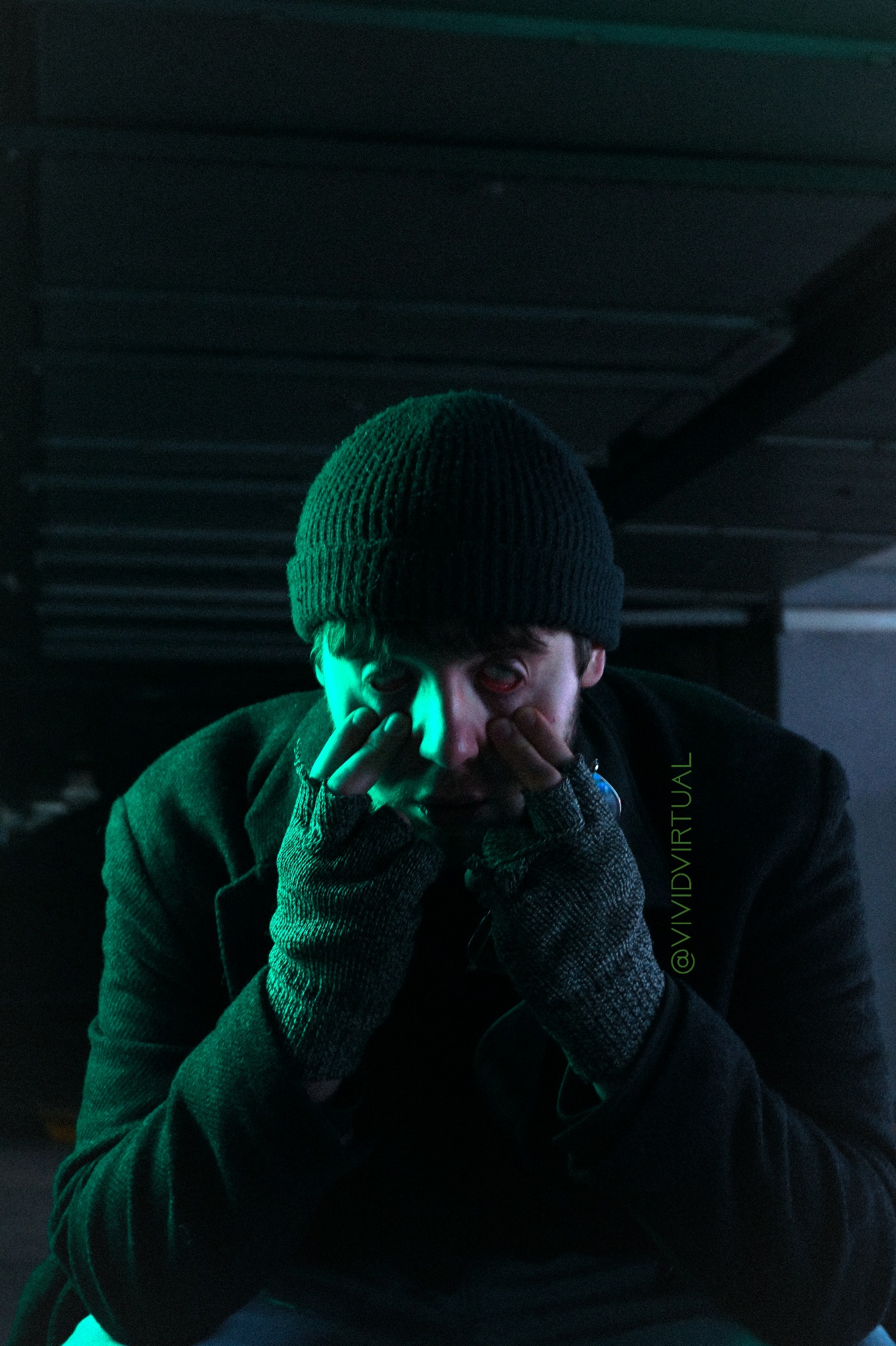 Mężczyzna w czapce i rękawiczkach, oświetlony zielonym światłem, dotyka twarzy dłońmi w rękawiczkach w ciemnym otoczeniu.