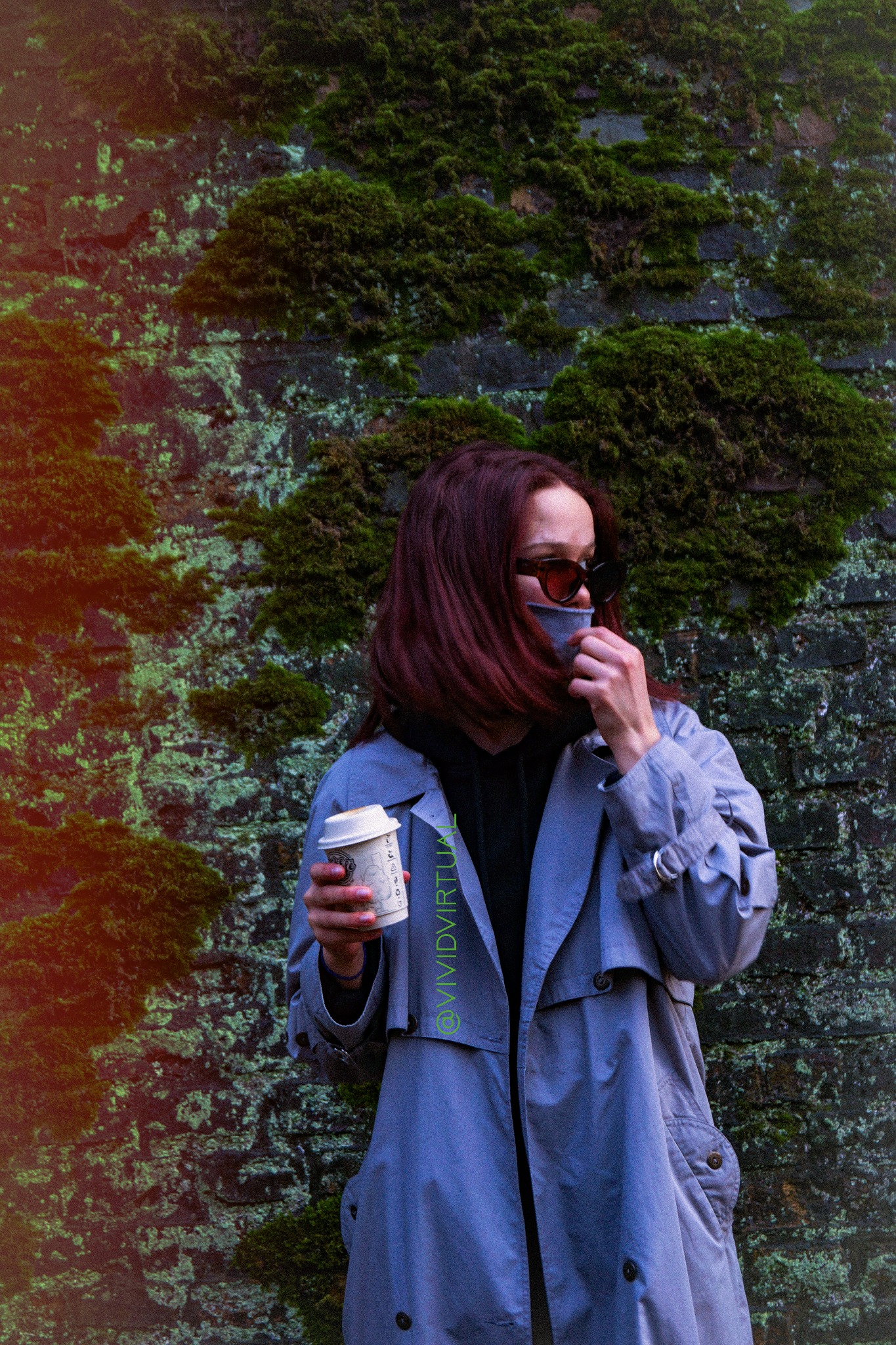 Kobieta w płaszczu i okularach przeciwsłonecznych, z kubkiem kawy, pozuje na tle ceglanej ściany porośniętej mchem w Poznaniu.