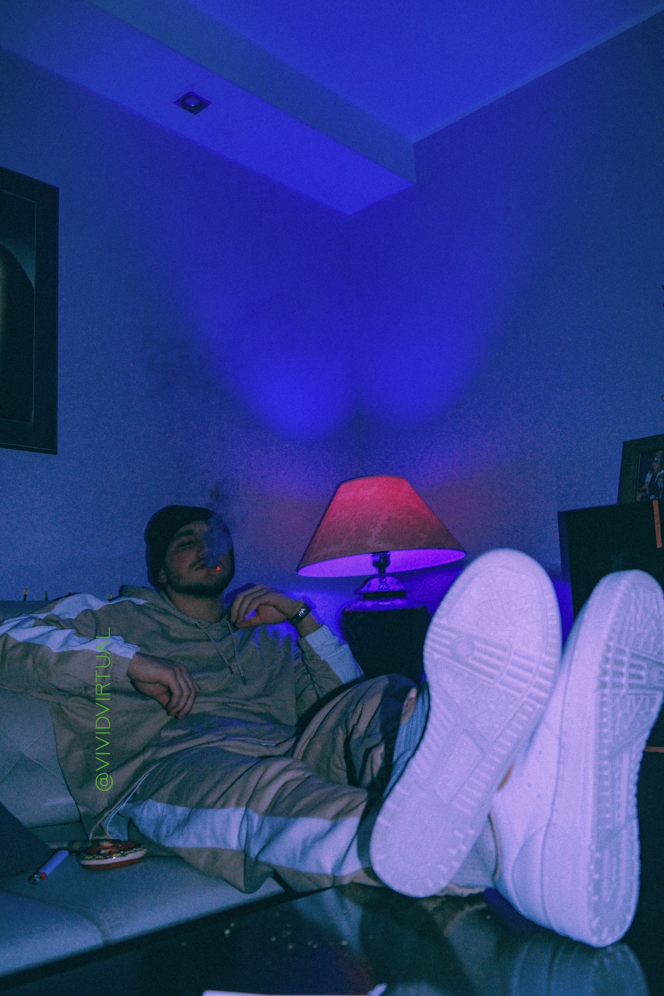 Mężczyzna w dresie leży na kanapie z uniesionymi nogami, paląc papierosa w pomieszczeniu oświetlonym niebieskim światłem, widoczne podeszwy butów i stolik.