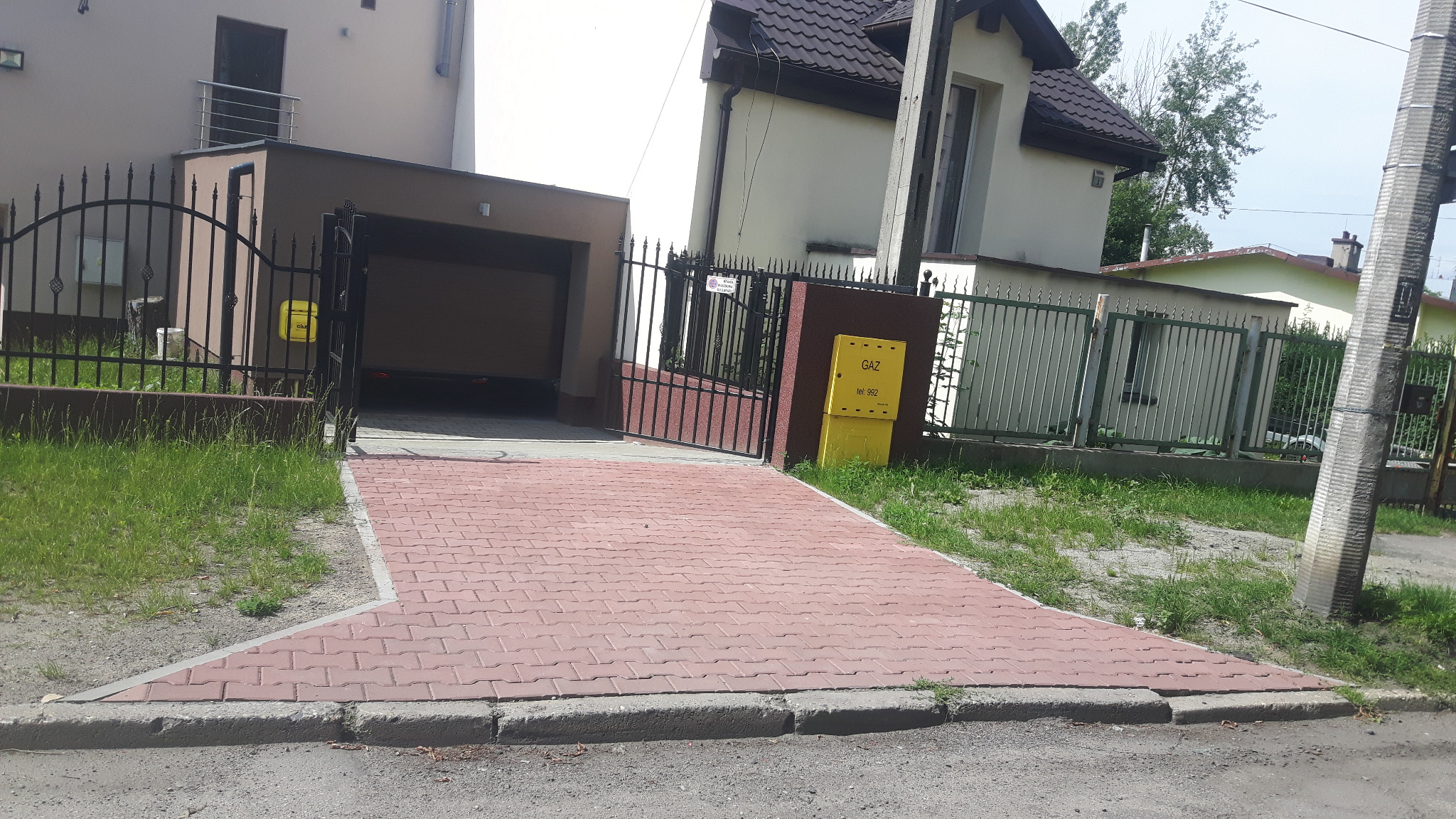 Podjazd z czerwonej kostki brukowej prowadzący do garażu w domu jednorodzinnym, widoczna skrzynka gazowa i ogrodzenie.