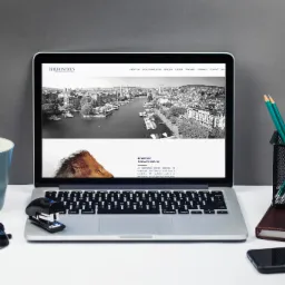 Laptop wyświetlający stronę internetową z panoramą miasta i wizerunkiem lwa, obok kubek, roślina, okulary i zszywacz.