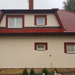 Dom jednorodzinny z beżową elewacją, czerwoną dachówką i obramowaniami okien w kolorze czerwonym, ozdobiony lampkami zwisającymi z dachu.