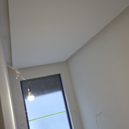 Wnętrze po malowaniu: białe ściany, sufit i okno zabezpieczone folią. Widoczne wystające kable i żarówka. Ujęcie z dołu, podkreślające świeżość wykończenia.