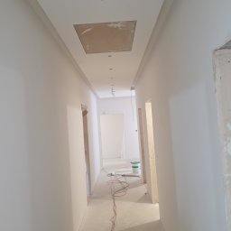 Korytarz po malowaniu na biało, widoczne gładkie ściany, sufit z otworem rewizyjnym i instalacją elektryczną. Na podłodze leżą kable, w tle wiadro z farbą.