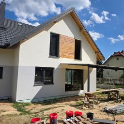 Dominik Salata - Dwukondygnacyjny dom z szarą dachówką i jasną elewacją w trakcie prac wykończeniowych. Widoczne rusztowania, narzędzia i materiały budowlane. Błękitne niebo w tle.