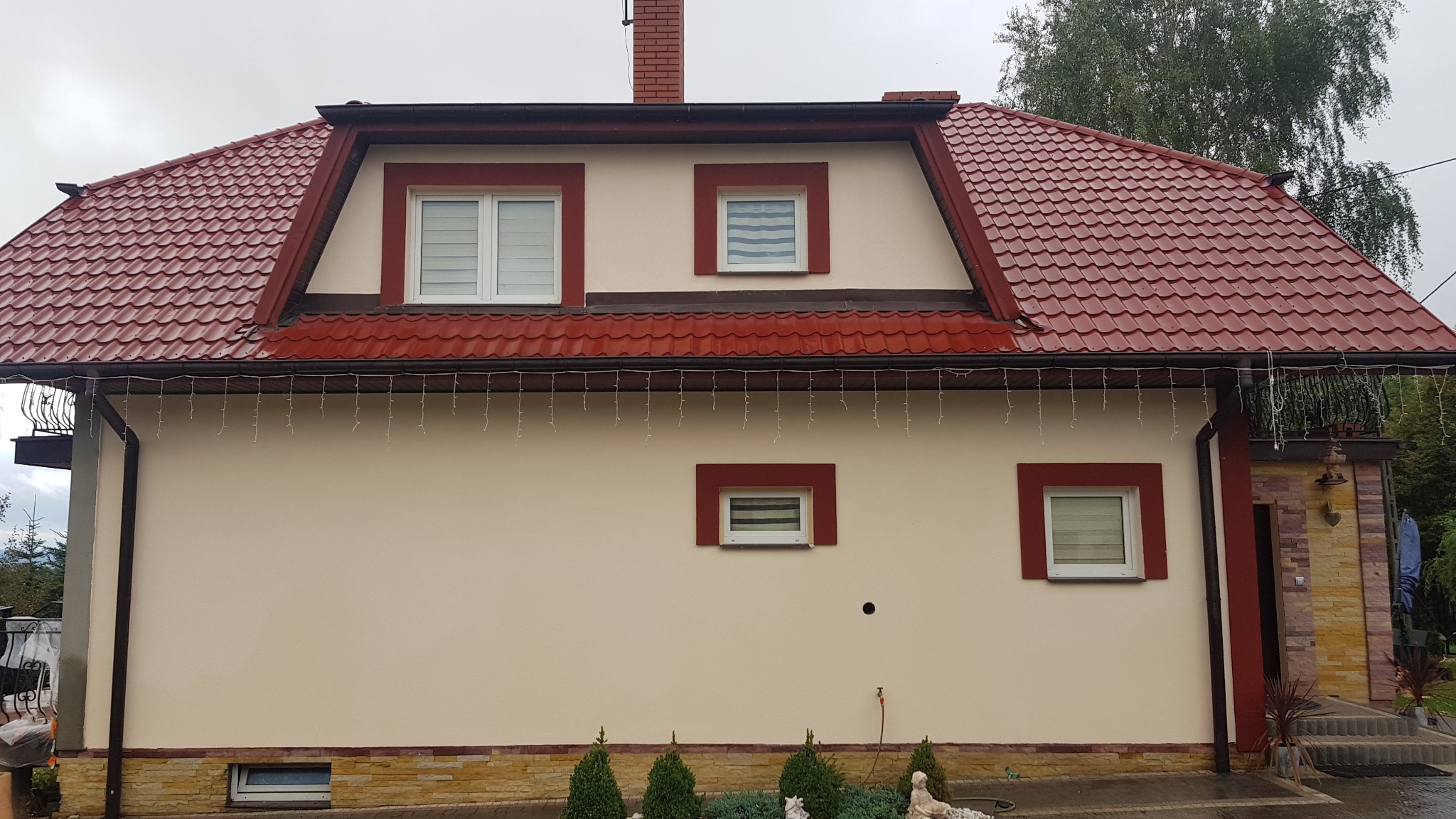 Dom jednorodzinny z beżową elewacją, czerwoną dachówką i obramowaniami okien w kolorze czerwonym, ozdobiony lampkami zwisającymi z dachu.