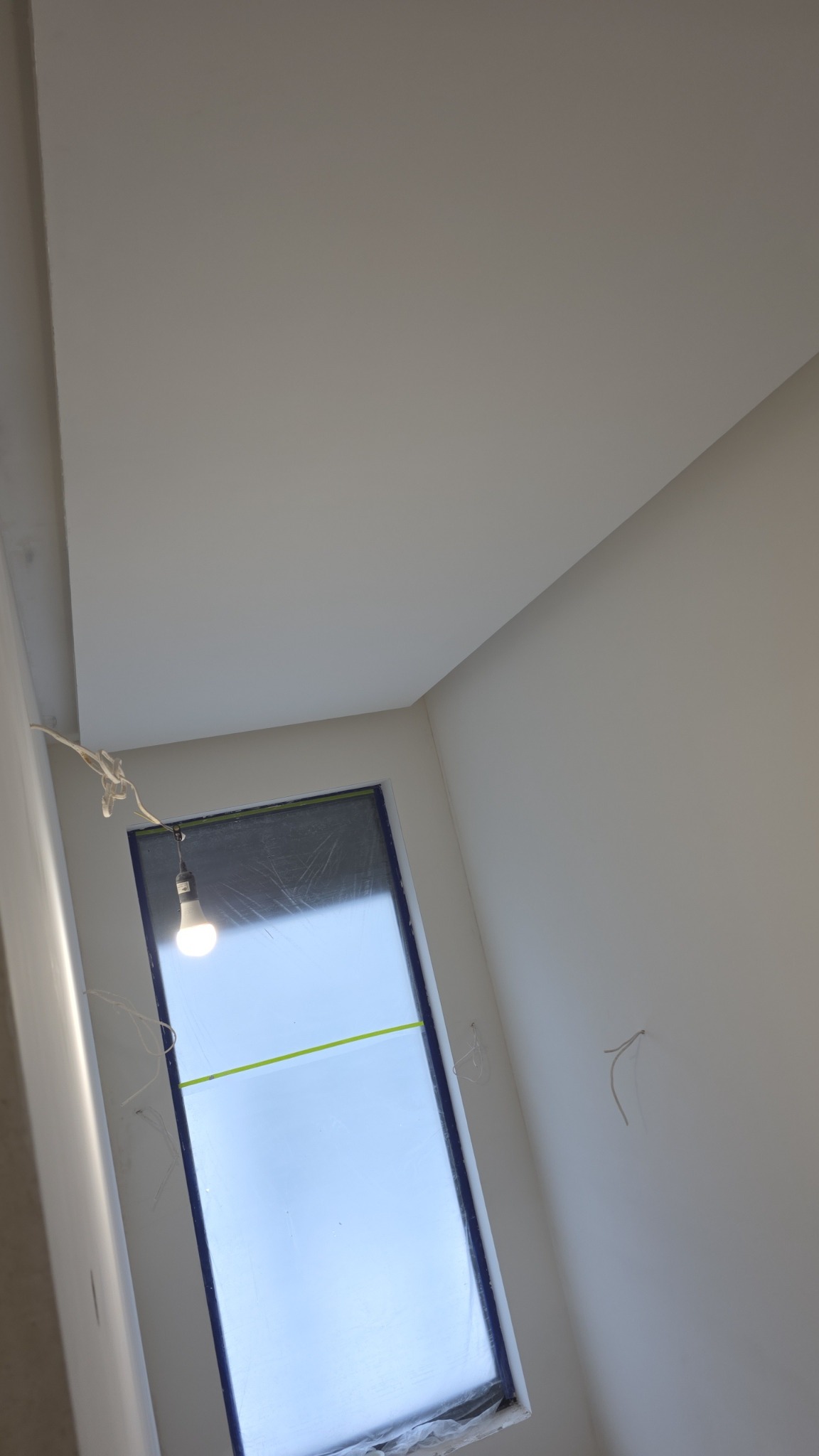 Wnętrze po malowaniu: białe ściany, sufit i okno zabezpieczone folią. Widoczne wystające kable i żarówka. Ujęcie z dołu, podkreślające świeżość wykończenia.