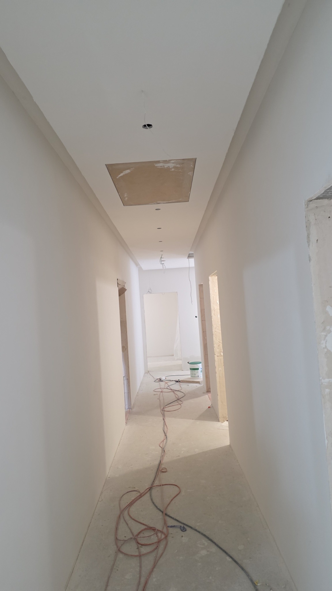 Korytarz po malowaniu na biało, widoczne gładkie ściany, sufit z otworem rewizyjnym i instalacją elektryczną. Na podłodze leżą kable, w tle wiadro z farbą.